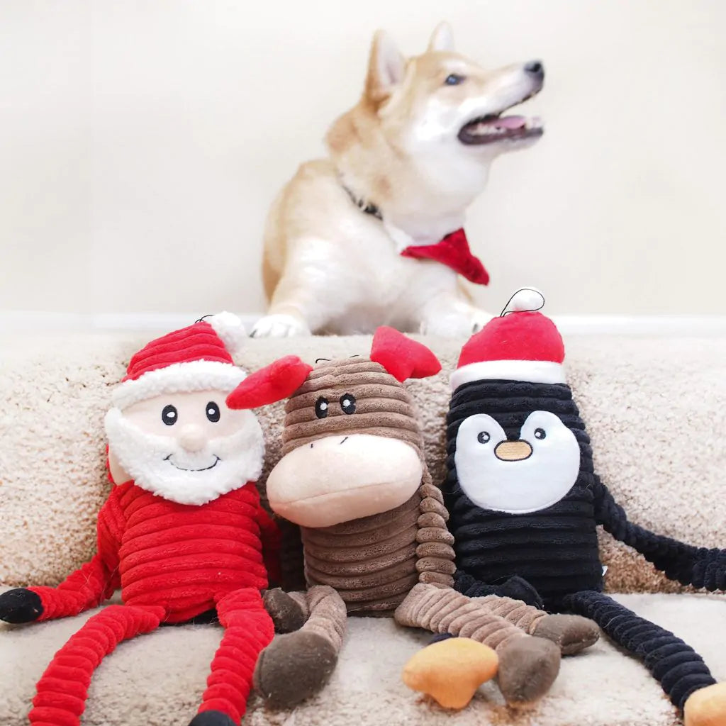 Three ZippyPaws Holiday Crinkle Dog Toys - Santa Claus, Moose and Penguin on a couch with a smiling Shiba Inu sitting behind them